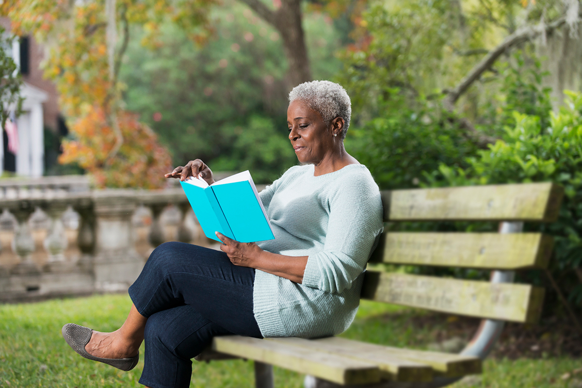 Senhora concentrada, sentada em um branco de praça, lendo um livro para mulheres empreendedoras