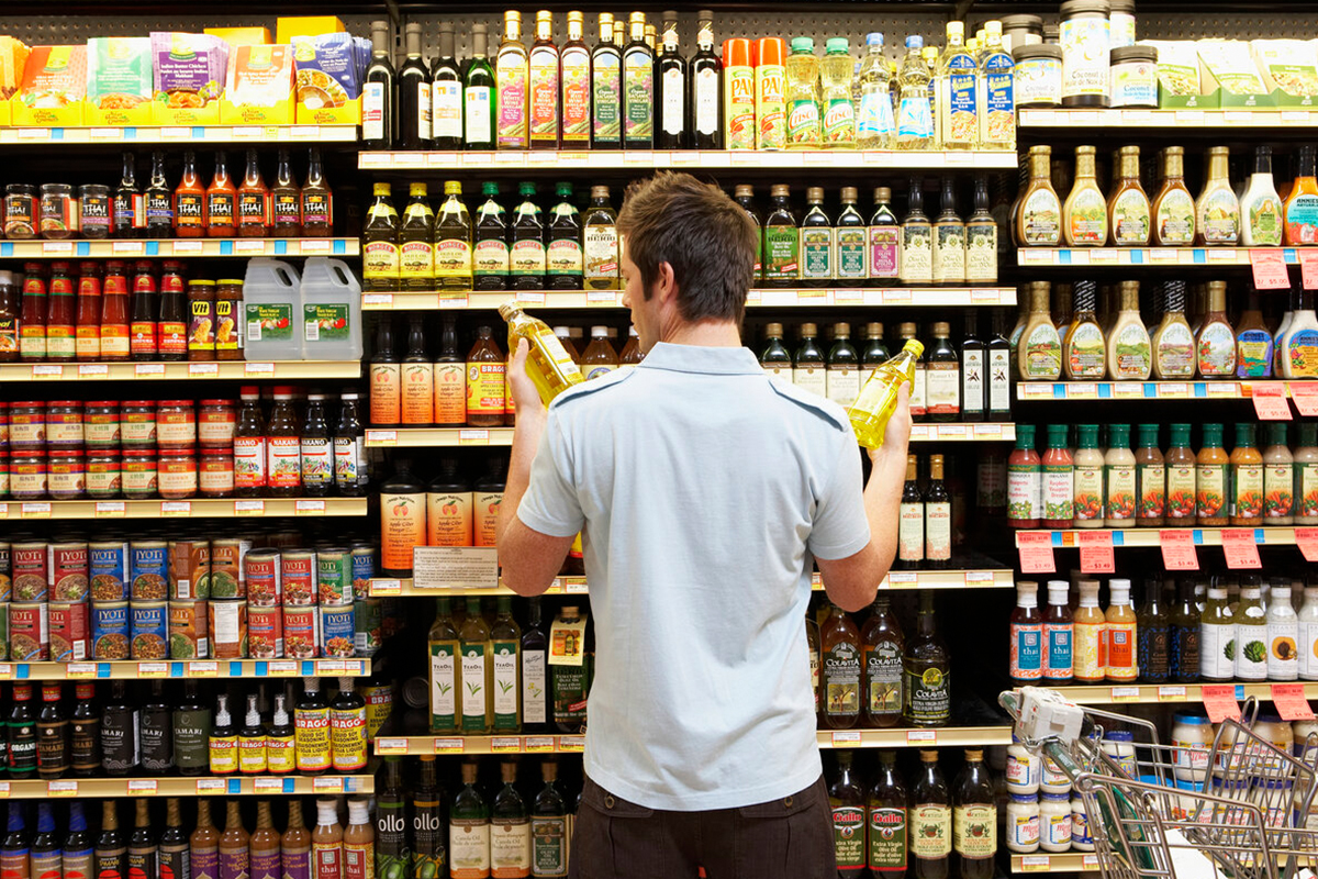 Homem pensativo, olhando para dois produtos no supermercado. Representa o upsell nas compras